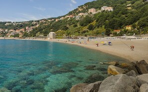 A coastal beach with clear water, rocky shoreline, and buildings on a hillside in the background. - [Dual Suite] Beaches 600m • Cruise Terminal 3km (Savona)
