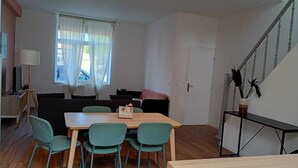A room with a wooden dining table, four chairs, a sofa, a TV on a stand, a window with curtains, and a staircase.