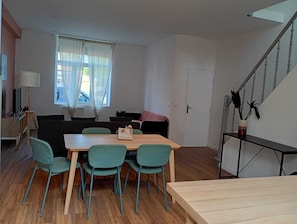 Huoneessa on puinen ruokapöytä, neljä tuolia, sohva, jalustalla oleva televisio, ikkuna, jossa on verhot, ja portaikko.