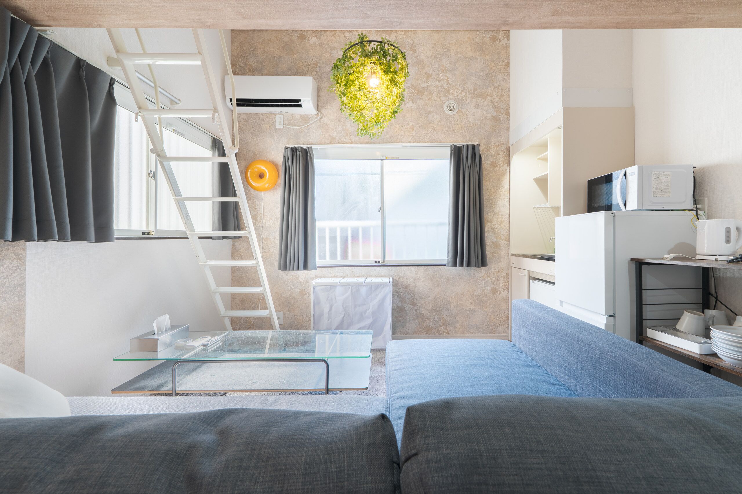 Um espaço de estar moderno com cama beliche, mesa de centro de vidro, sofá, cozinha e planta pendente.