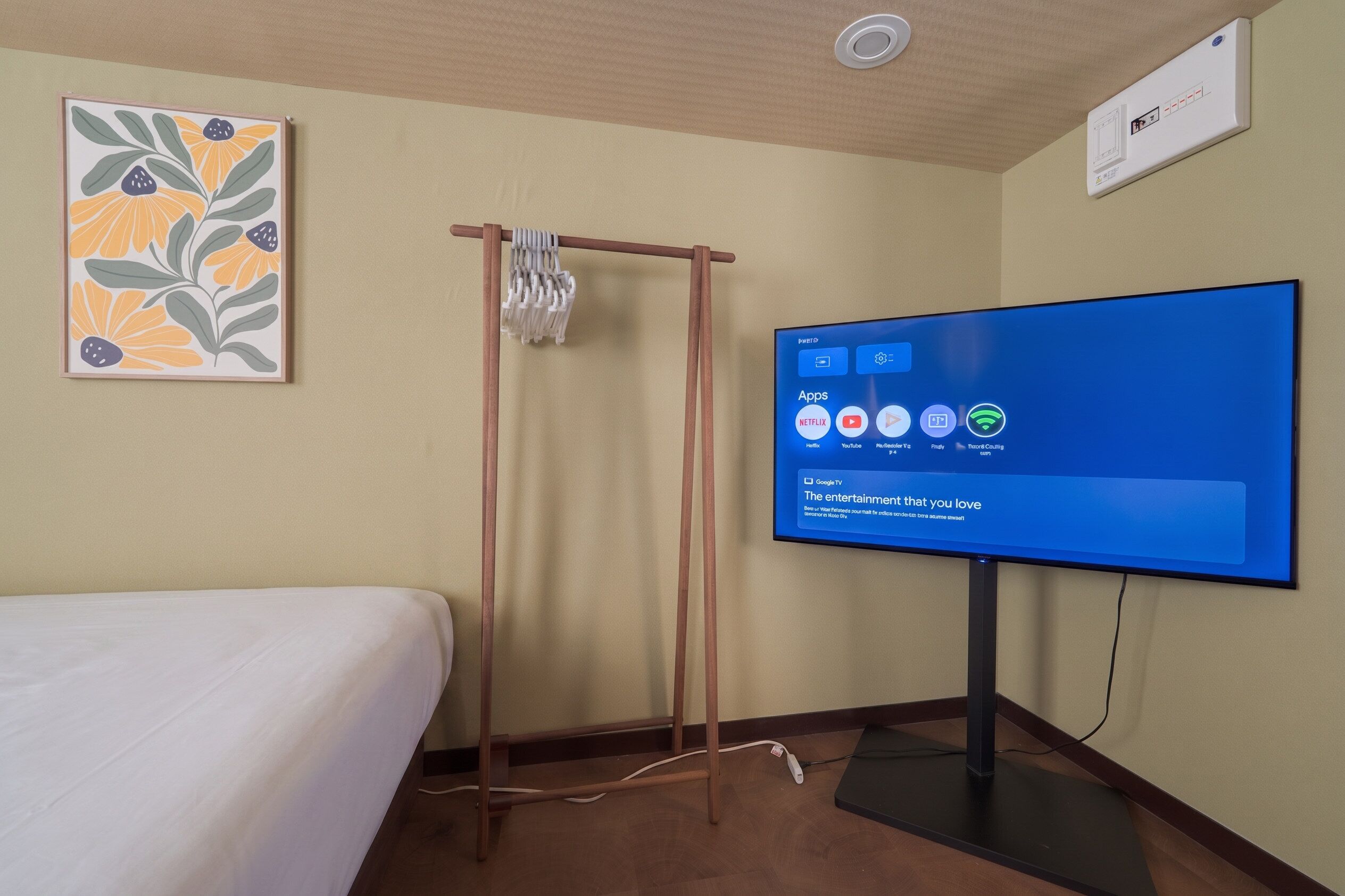 A hotel room with a bed, a TV displaying entertainment apps, a wall-mounted air conditioner, and a framed floral artwork.