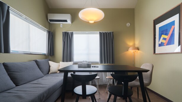 A compact living space with a sofa, a dining table with chairs, an air conditioning unit, and a lamp.