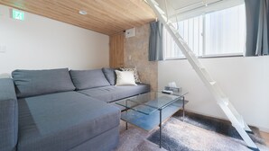 A modern living room with a grey sectional sofa, a glass coffee table, a wooden ceiling, a staircase, and a window with curtains.