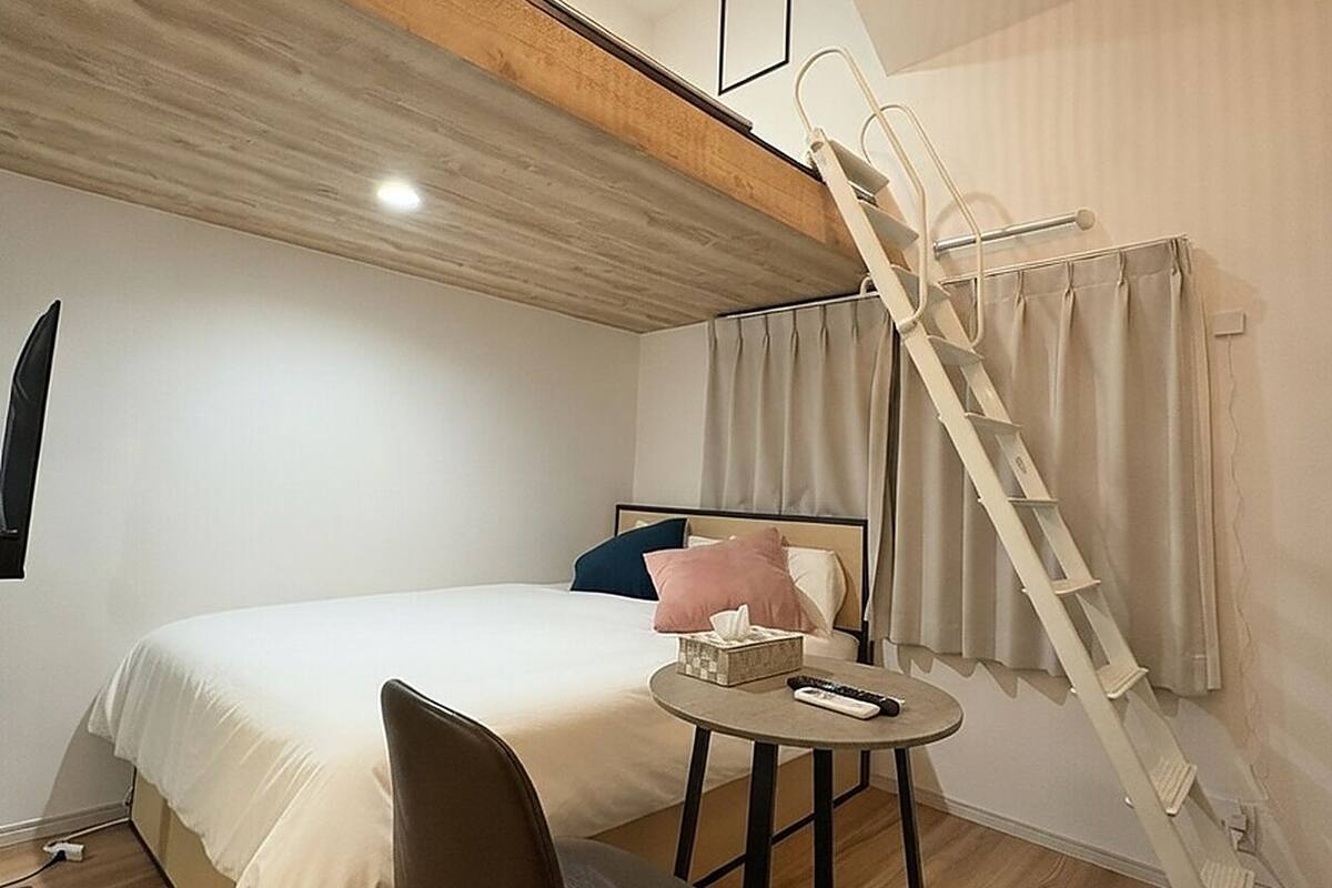 A bedroom with a bed, a bedside table with a remote control, a chair, a ladder, and a window with curtains.