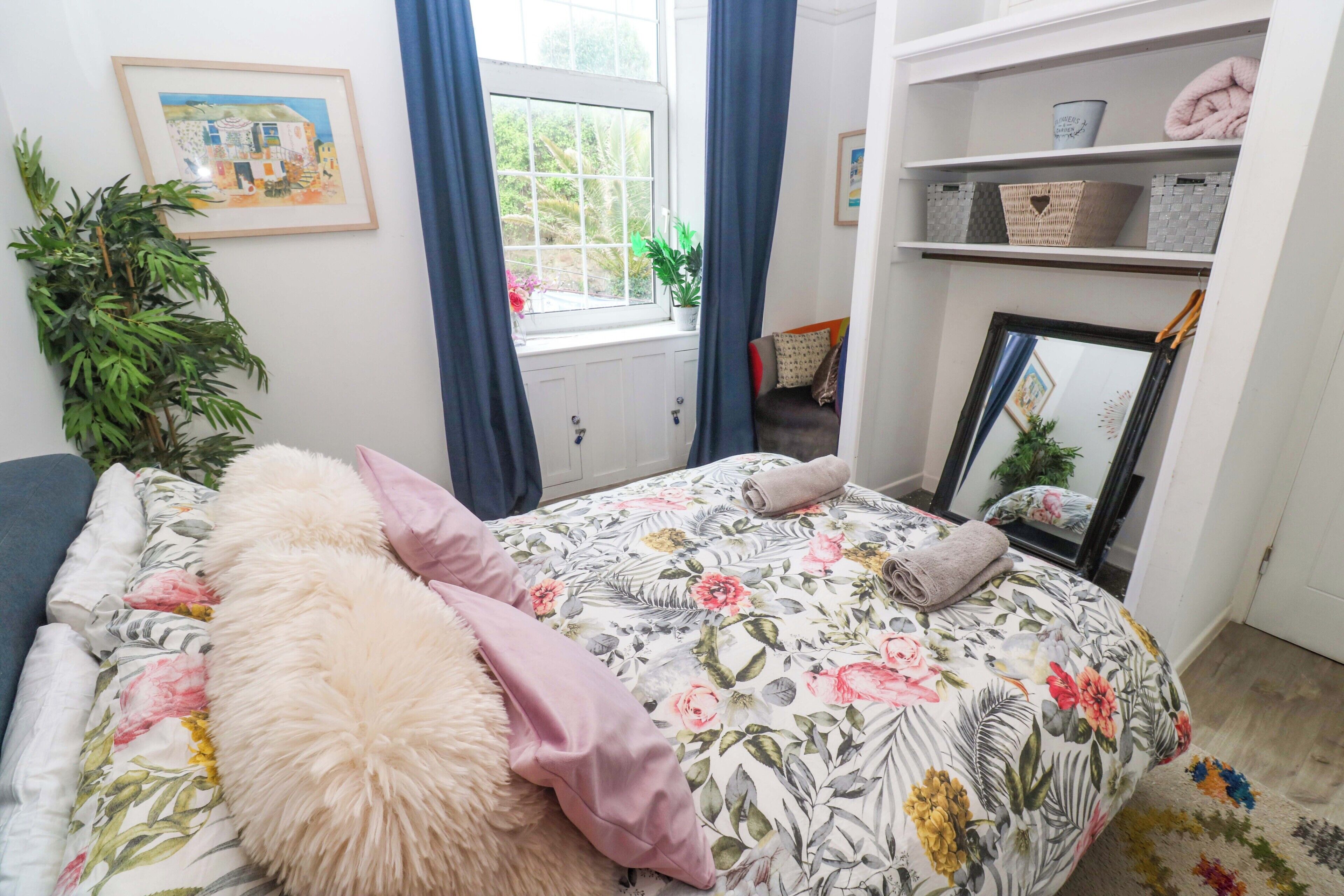 Quarto com colcha florida, uma planta grande em vaso, janela com cortinas e prateleira com cestos organizadores.