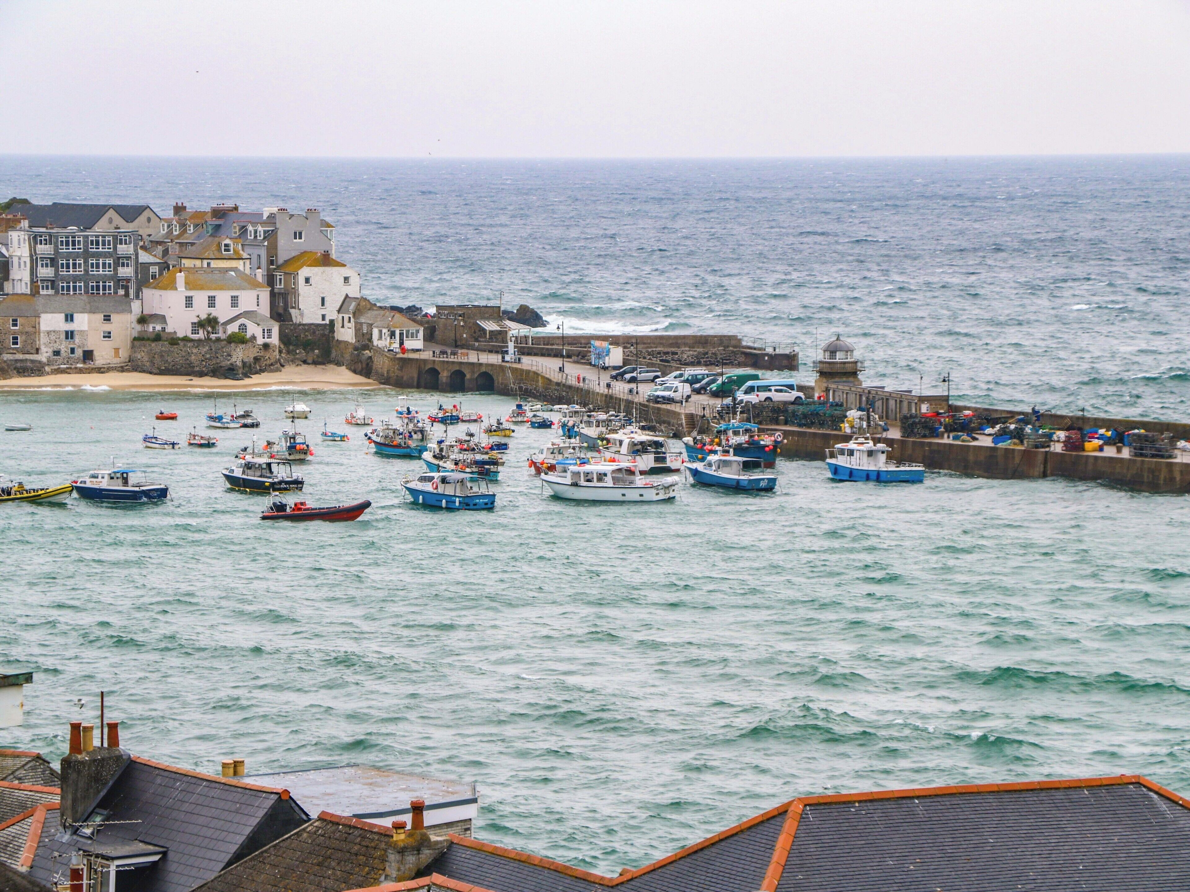 A coastal town with a harbor filled with boats, a lighthouse, and buildings along the shore.