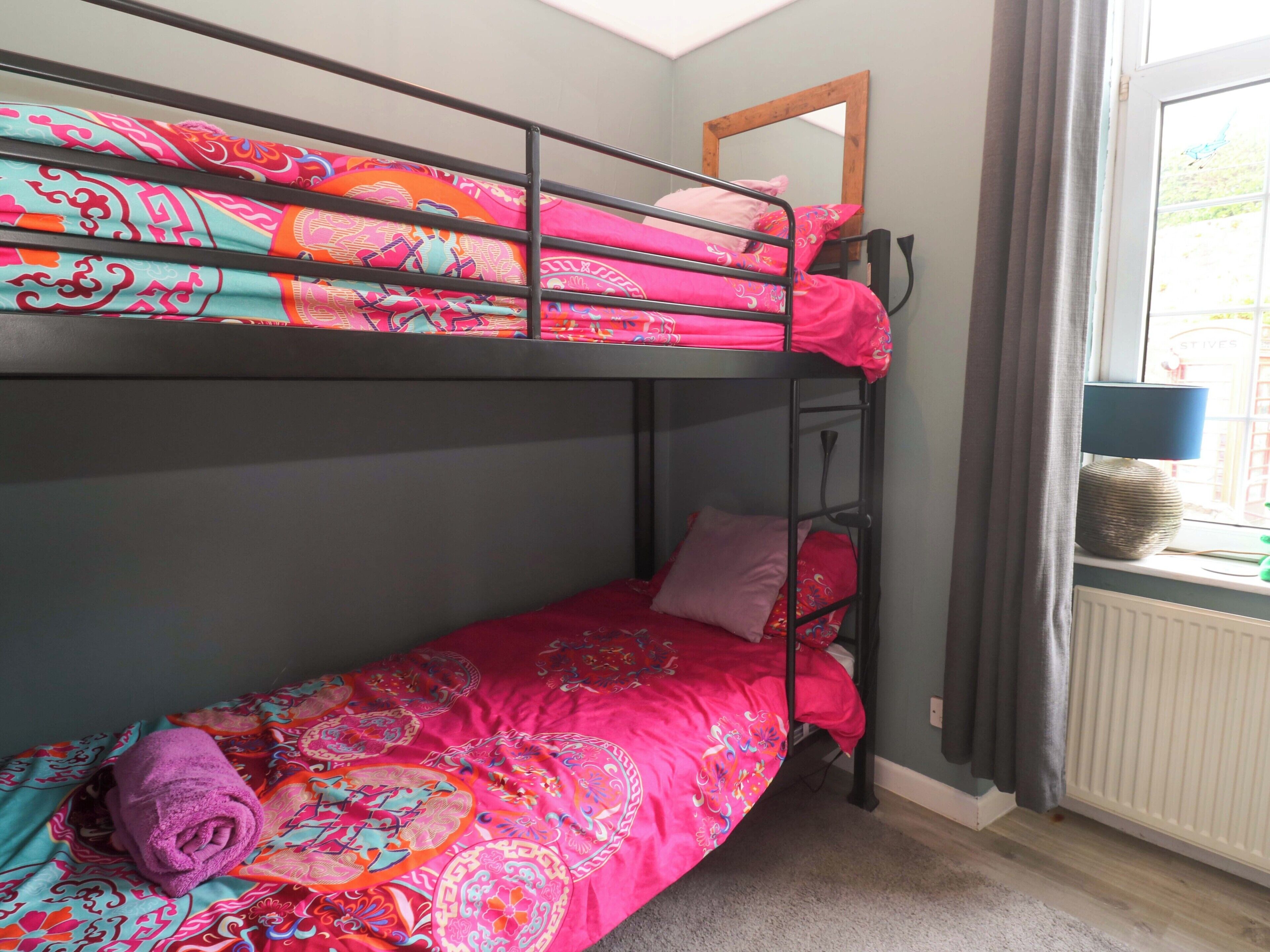 A bunk bed with colorful bedding, a window with curtains, and a lamp on a bedside table.