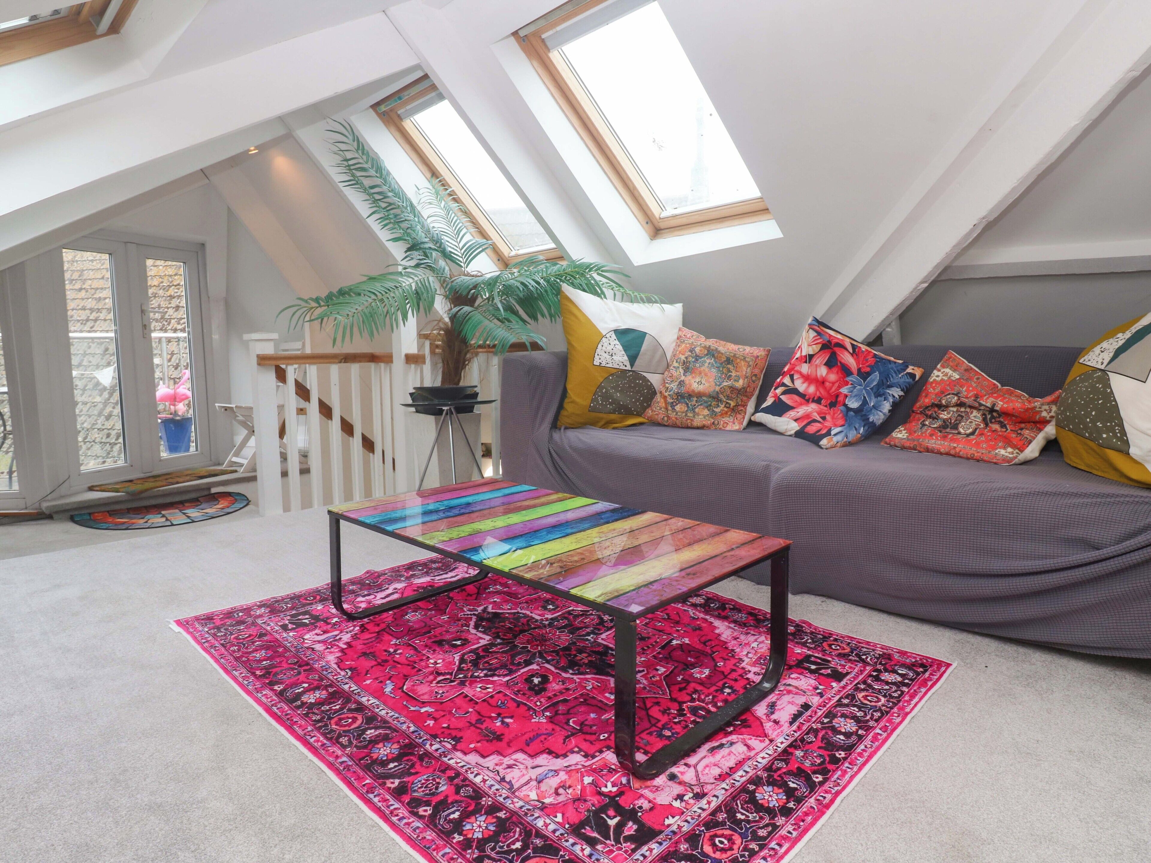 A loft-style room with a sloped ceiling, a grey sofa with colorful cushions, a glass coffee table, a red patterned rug, and a potted plant.