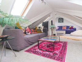Una habitación tipo loft con techo inclinado, un sofá gris con cojines coloridos, una mesa de centro de vidrio, un tapete estampado y una planta grande.