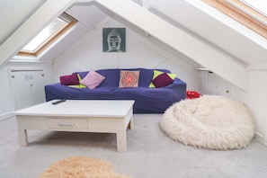 Un dormitorio tipo loft con techo inclinado, un sofá azul con cojines estampados, un escritorio blanco, un otomán mullido y un cuadro de Buda.