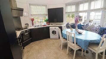 A compact kitchen with a stove, oven, and washing machine, a dining area with a table and chairs, and large windows with blinds.