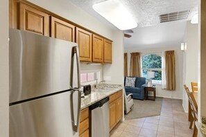 A kitchen with wooden cabinets, a stainless steel refrigerator, and a dishwasher. There is a window with curtains and a view of greenery outside. - Cozy 2-bedroom apartment in fantastic St. George with AC and gym Southgate (St. George)