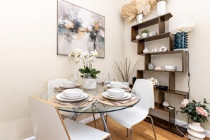 A dining area with a glass table, white chairs, and a modern abstract painting on the wall. - Comfortable Apartments in Notting Hill + Aircon,  2m, Walk to Train & Hyde Park (London)