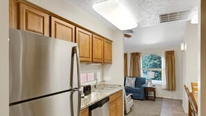 A kitchen with wooden cabinets, a stainless steel refrigerator, and a dishwasher. There is a window with curtains and a view of greenery outside. - Cozy 2-bedroom apartment in tranquil St. George with AC and gym Southgate (St. George)