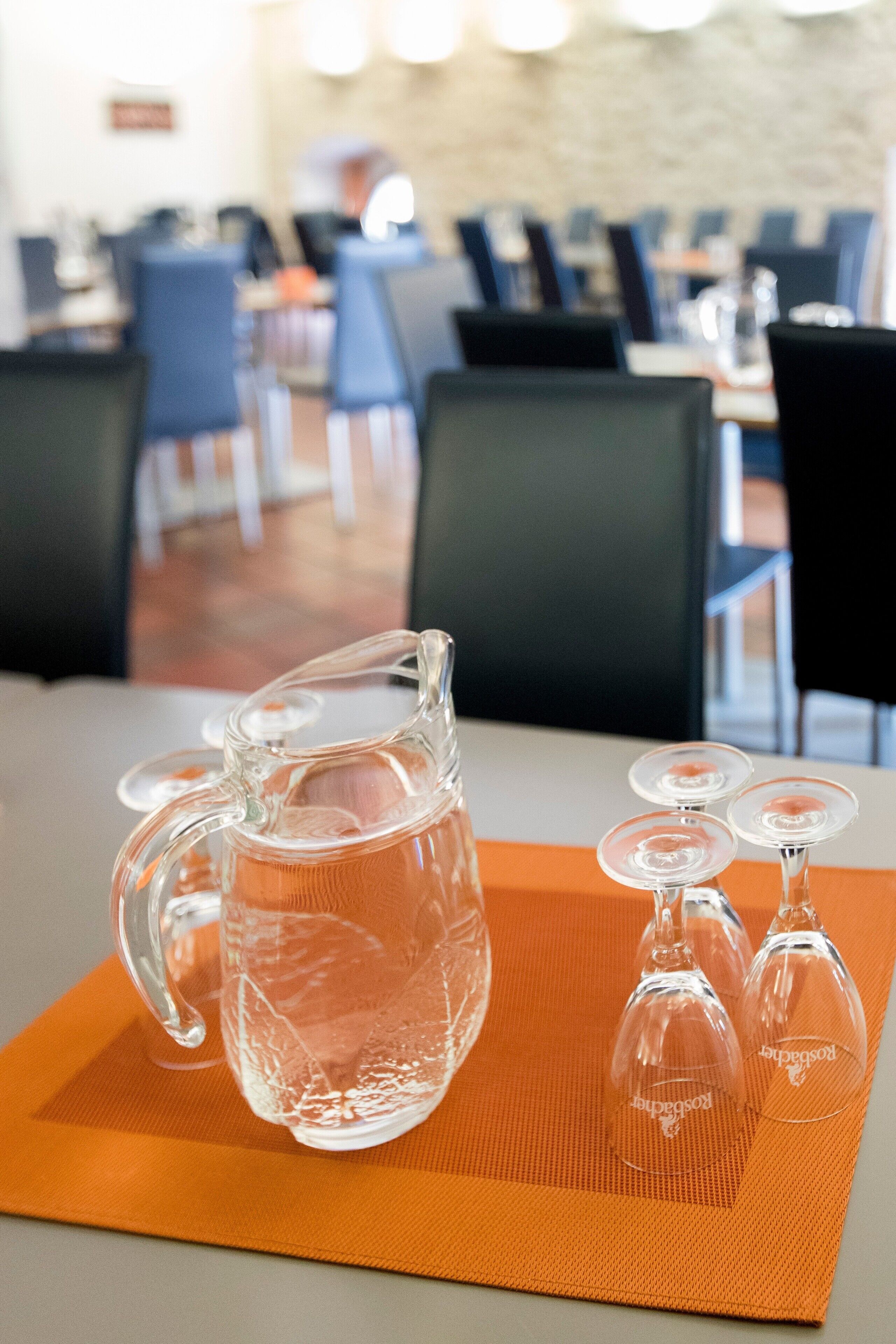 Une table avec une carafe et des verres à vin, le tout posé sur un set de table orange, dans un coin repas agrémenté de chaises et de tables.