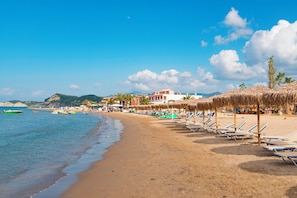 A beach with rows of sun loungers, a thatched-roof structure, and a building in the background. - Louvre Sidari Suite (Sidari)