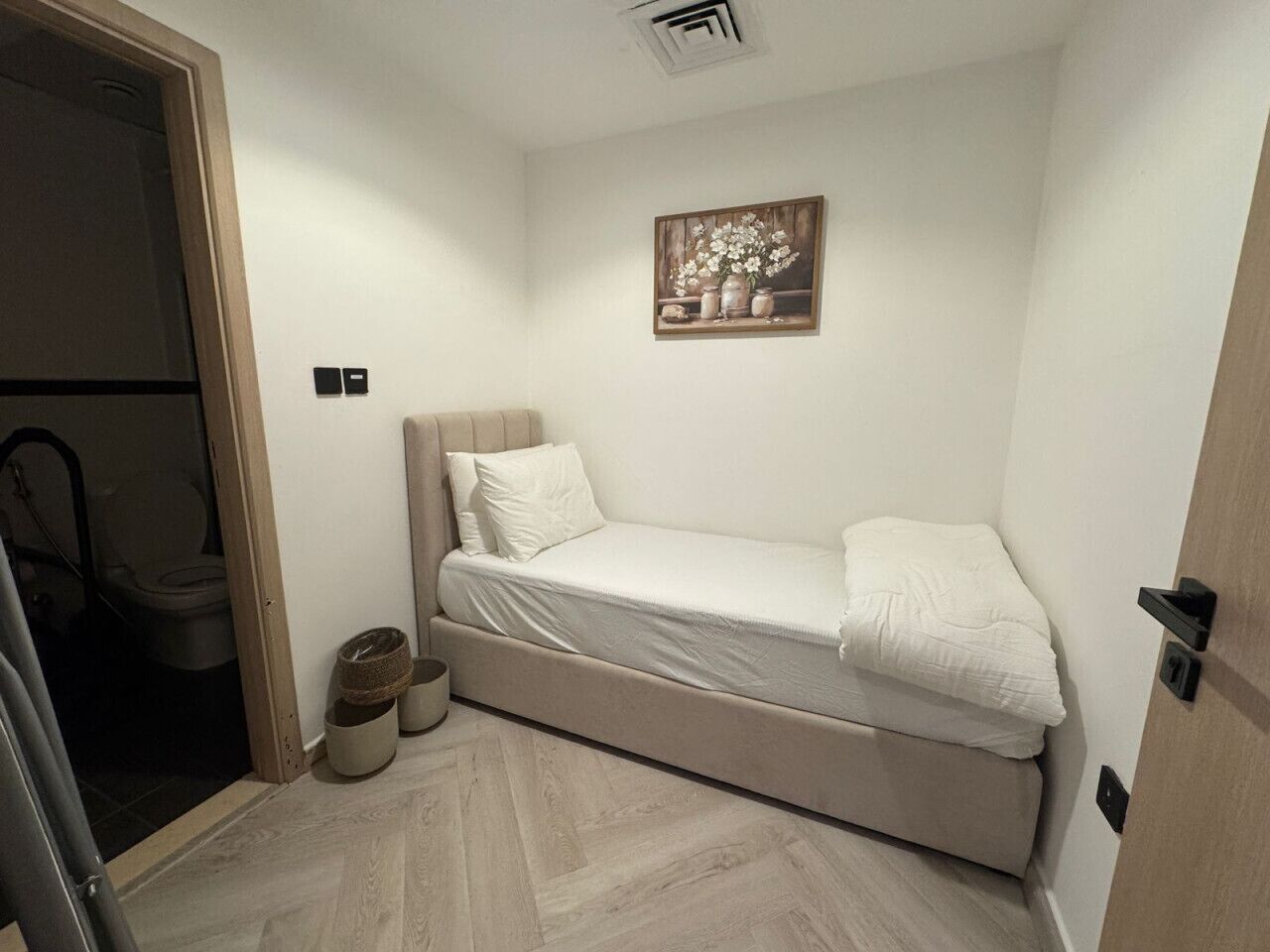 Ein kleines Zimmer mit einem Einzelbett, einer Holztür, einem kleinen Nachttisch und einem an der Wand hängenden Bild.