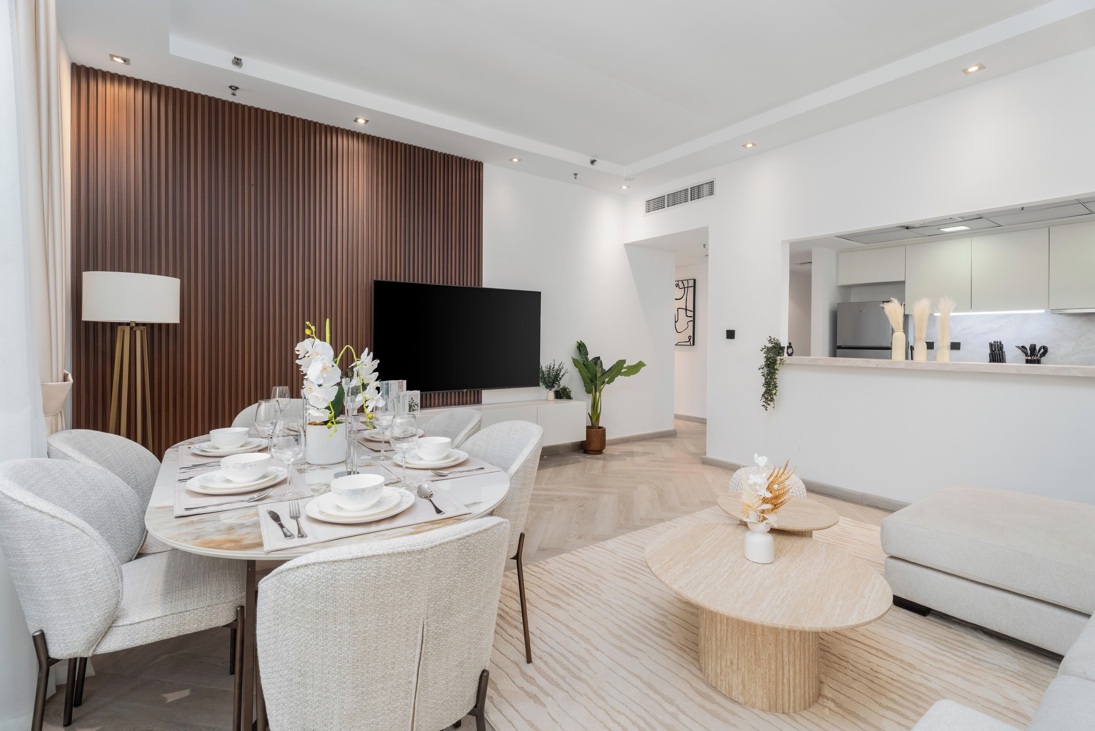Un espace repas moderne avec une table ronde dressée pour six personnes, un téléviseur à écran plat fixé au mur et une cuisine visible à travers une porte ouverte.
