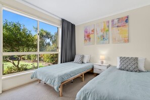 A bedroom with two beds, a window with curtains, a bedside table with a lamp, and abstract wall art. - The Happy Bay House (Apollo Bay)