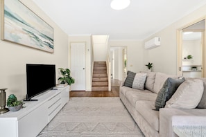 A modern living room with a flat-screen TV, a beige sofa, and a staircase leading to another level. - The Happy Bay House (Apollo Bay)