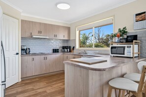 A modern kitchen with light wood cabinets, a central island, and a window providing a view of trees. - The Happy Bay House (Apollo Bay)