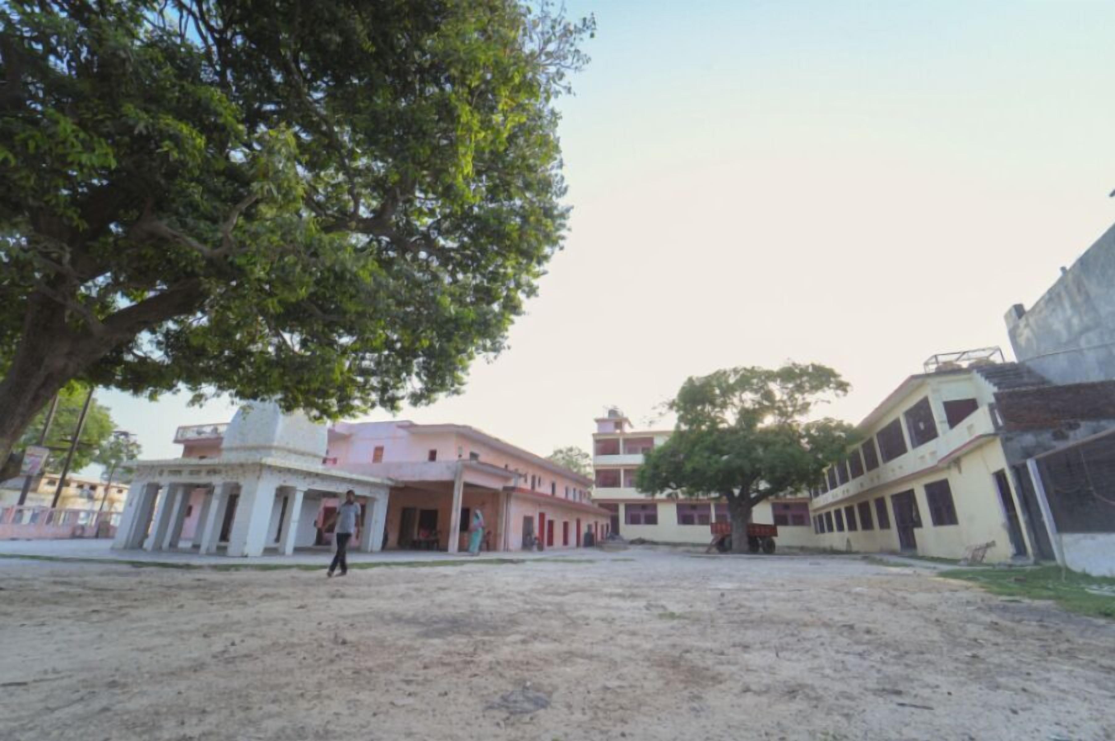 A building with multiple floors, a large tree in the foreground, and a dirt ground.