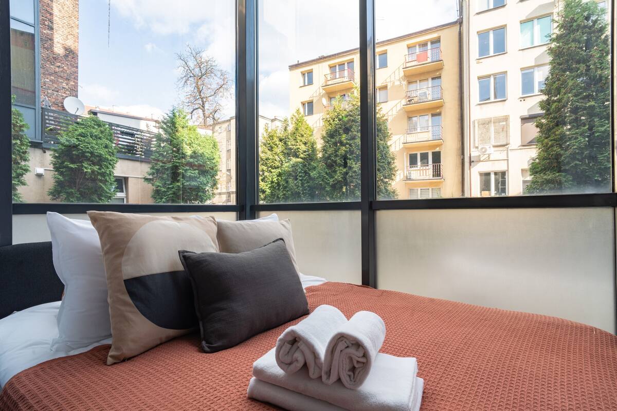 A hotel room with a bed, pillows, and a view of buildings and trees through large windows.