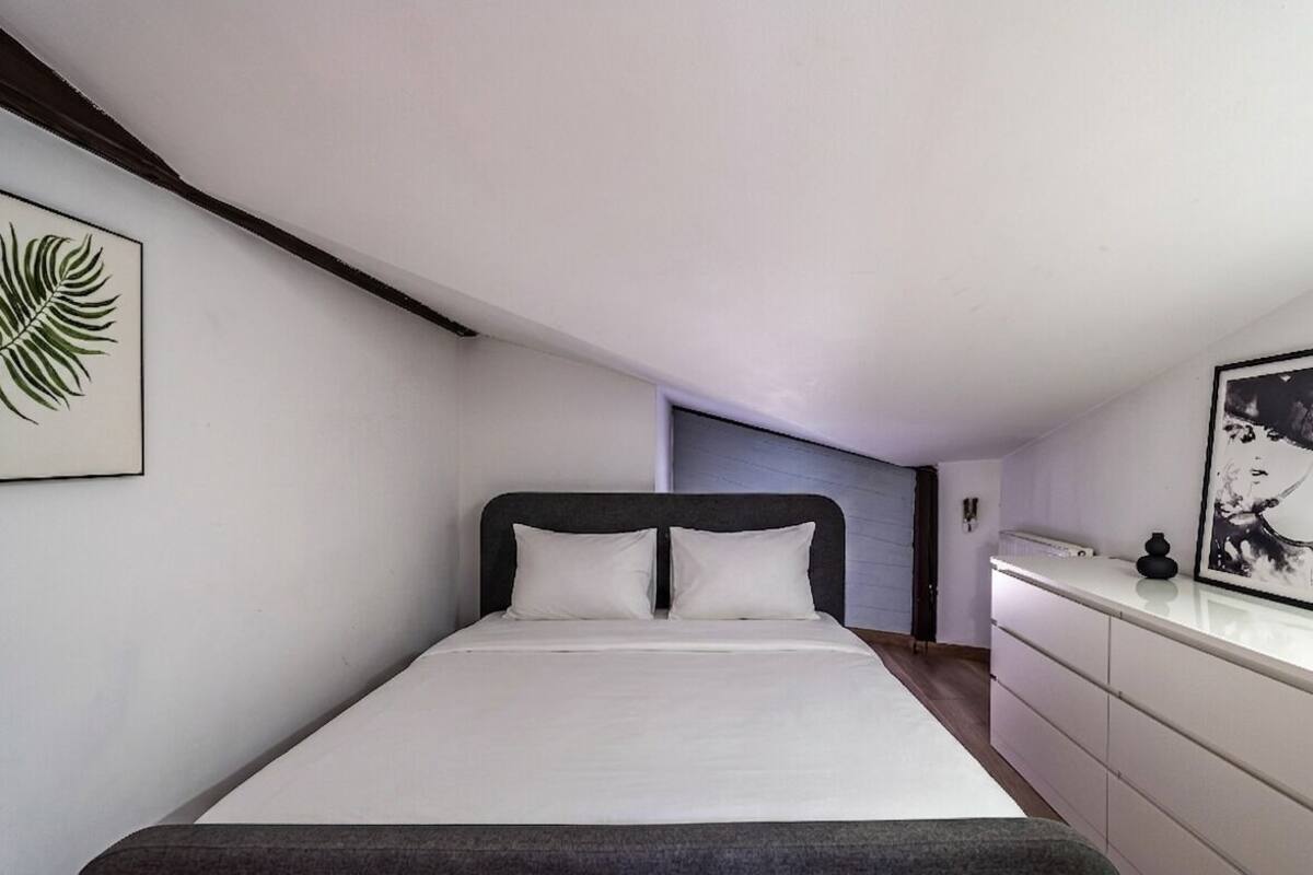 A bedroom with a bed, a nightstand, a framed picture on the wall, and a door leading to another room.