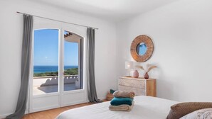 Une chambre avec un grand lit, une table de chevet, un miroir et une vue sur la mer, accessible par une porte coulissante.