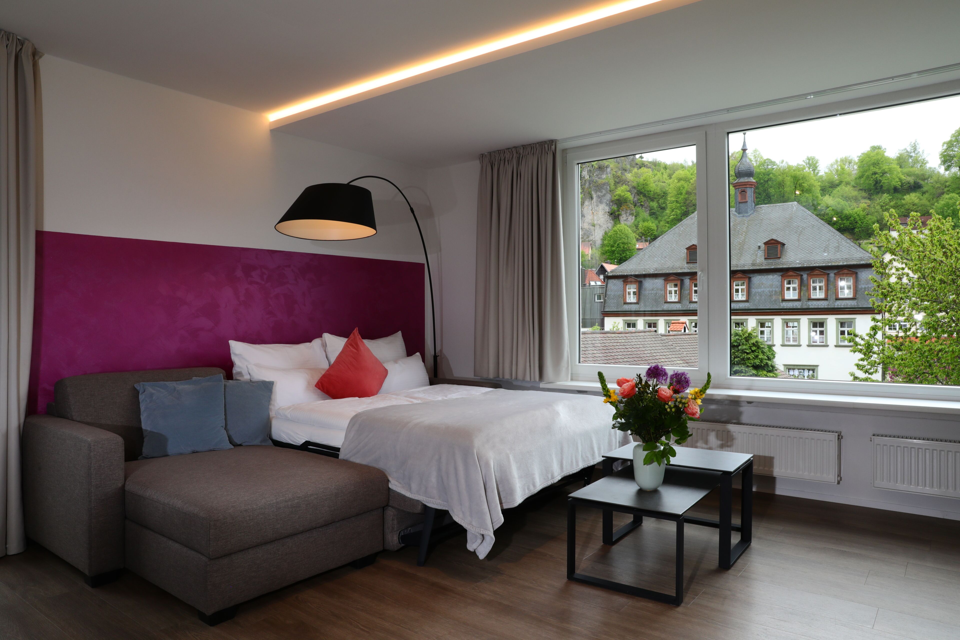Une chambre moderne avec un lit, un canapé, une petite table et une fenêtre donnant sur un immeuble.