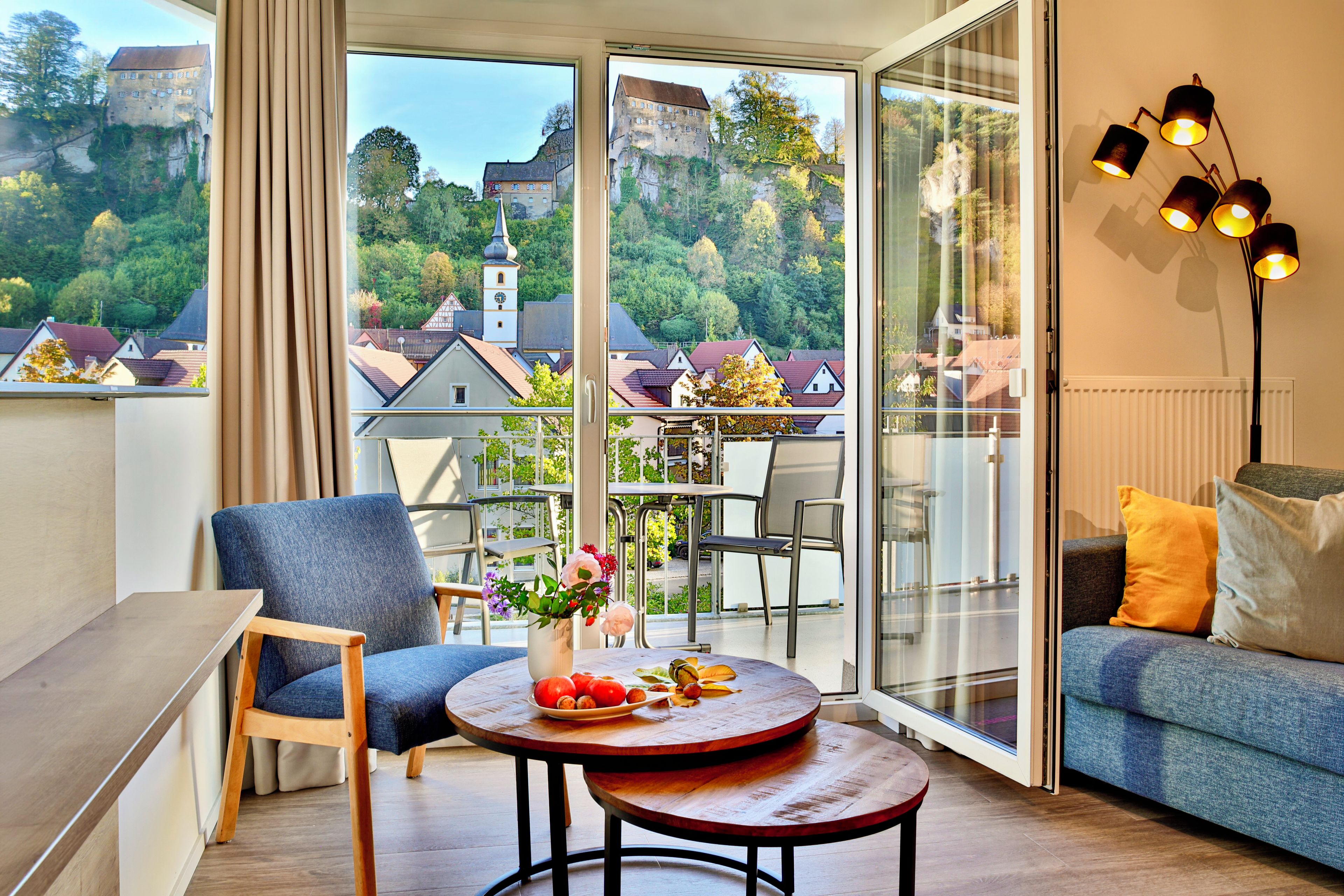 A modern living room with a sofa, a round table with a vase of flowers, and a chair. Large windows offer a view of a historic building and trees.