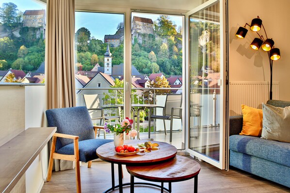 A modern living room with a sofa, a round table with a vase of flowers, and a chair. Large windows offer a view of a historic building and trees.