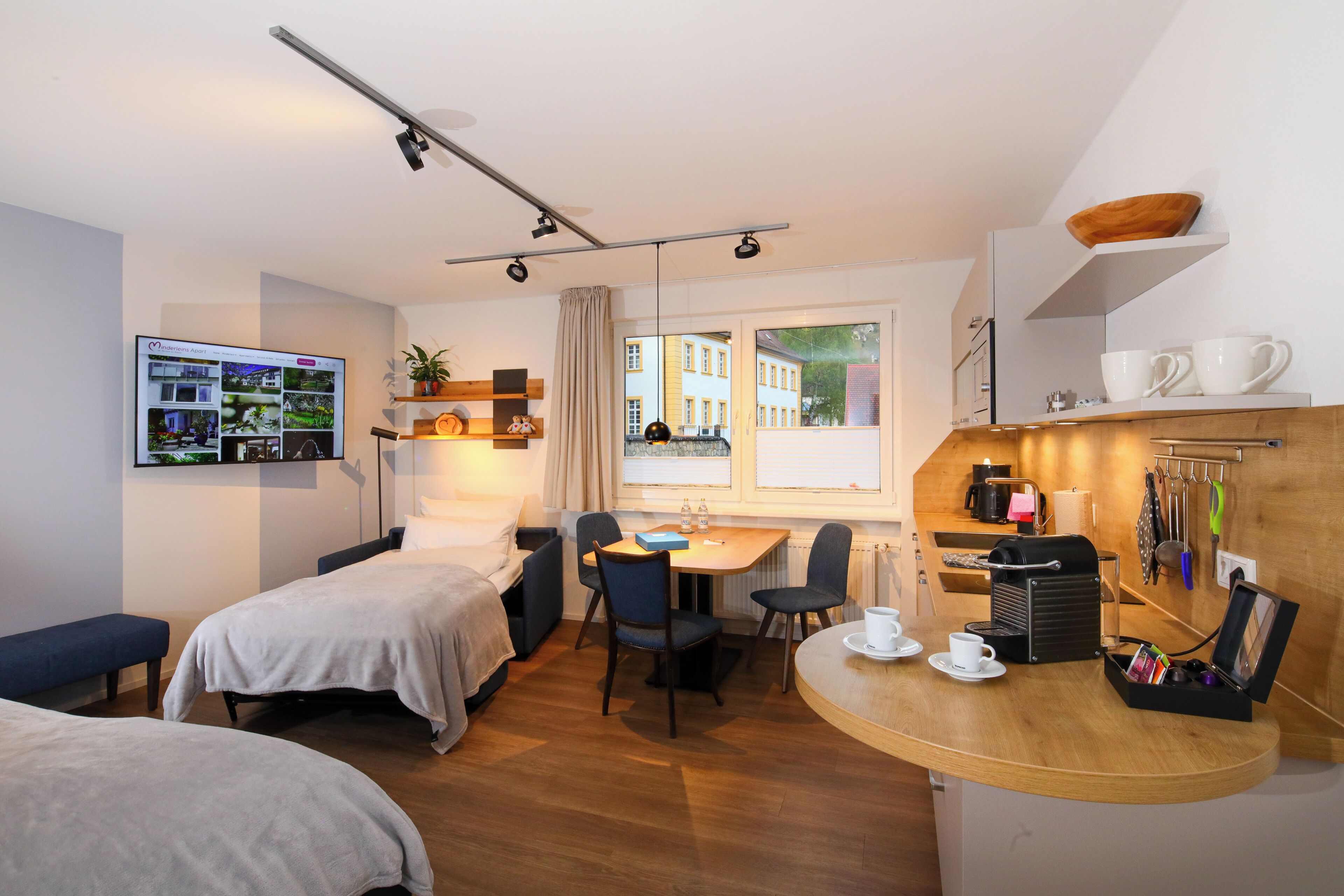 Habitación de hotel compacta con dos camas, una pequeña mesa de comedor, una cafetera y vistas a edificios a través de la ventana.