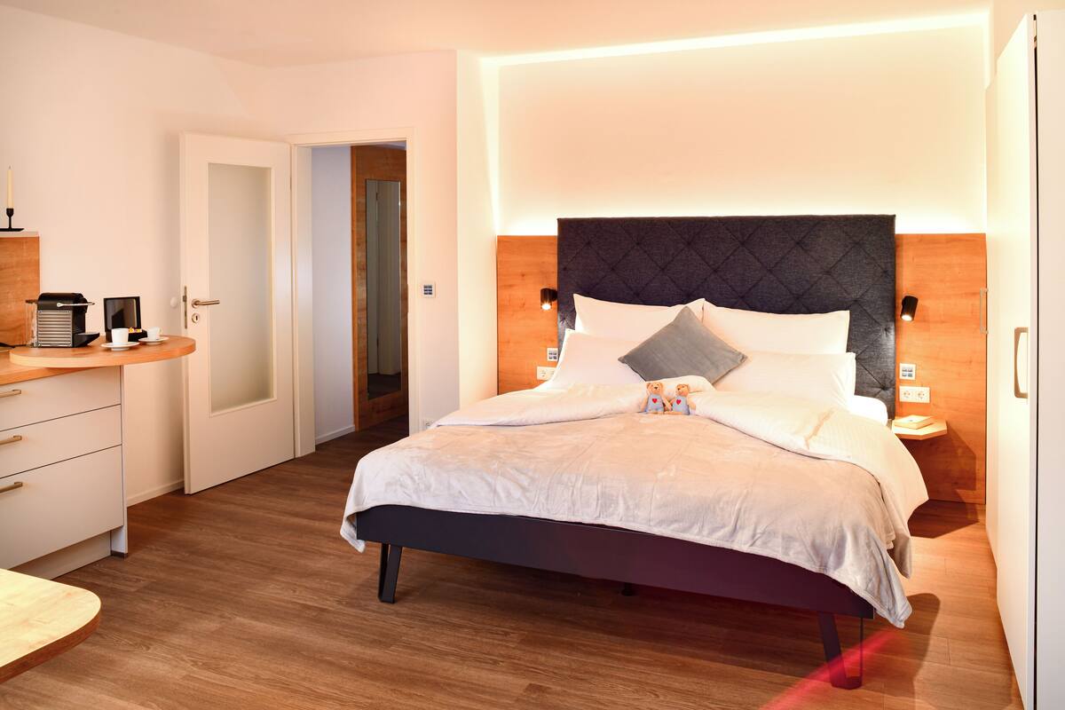 Una habitación de hotel moderna con una cama, mesitas de noche, un escritorio y una puerta que da a otra habitación.