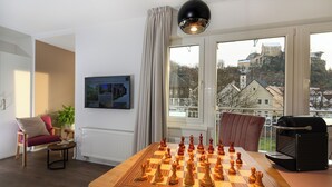 Una sala de estar con un tablero de ajedrez grande, un sofá, una mesa de centro, una planta en maceta y vistas de un castillo por la ventana.