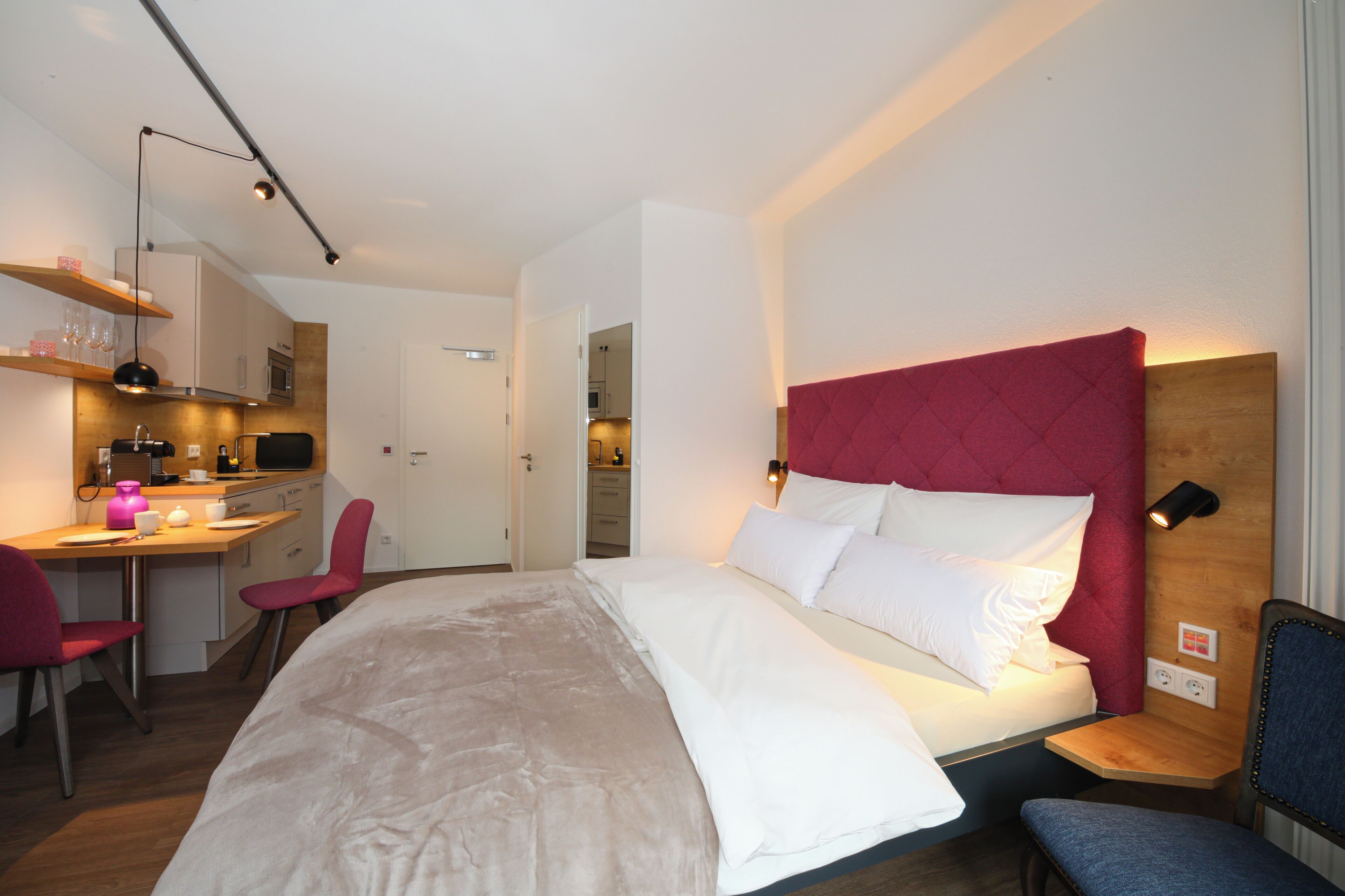Une chambre d’hôtel moderne, équipée d’un lit, d’un bureau avec une chaise, d’une kitchenette et d’une salle de bain visible à travers une porte ouverte.