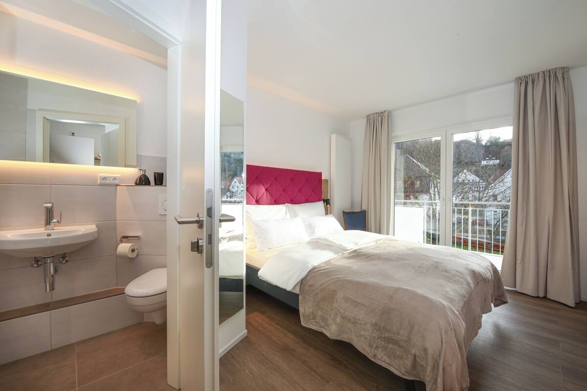 Una habitación de hotel moderna con baño, una cama con cabecera roja y vistas al exterior.