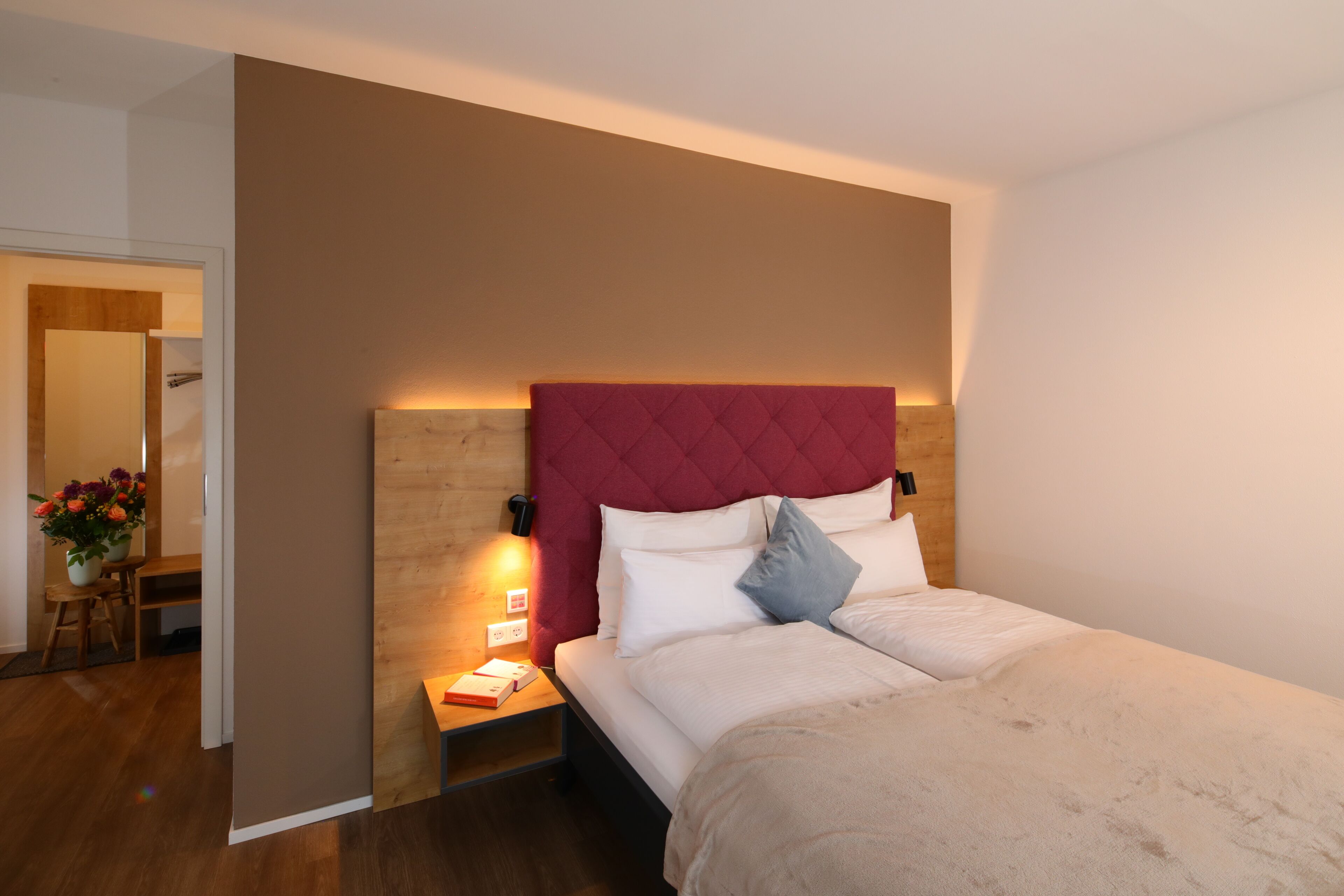 Un dormitorio con cabecera de madera, una mesita de noche con lámpara, cama con ropa de cama blanca y una pequeña mesa con un florero.