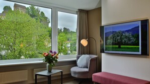 Una habitación con un ventanal grande, una silla, un banco, una mesa con un florero y un televisor en la pared.