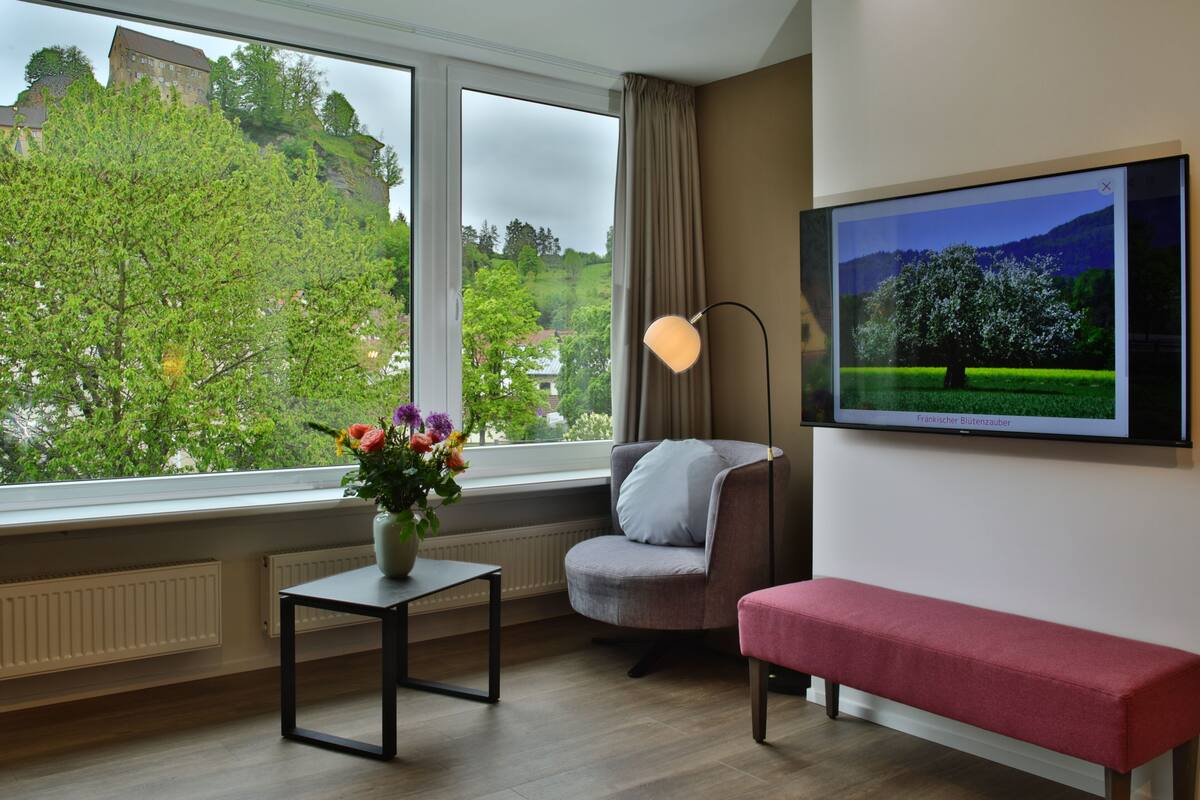 Una habitación con un ventanal grande, una silla, un banco, una mesa con un florero y un televisor en la pared.