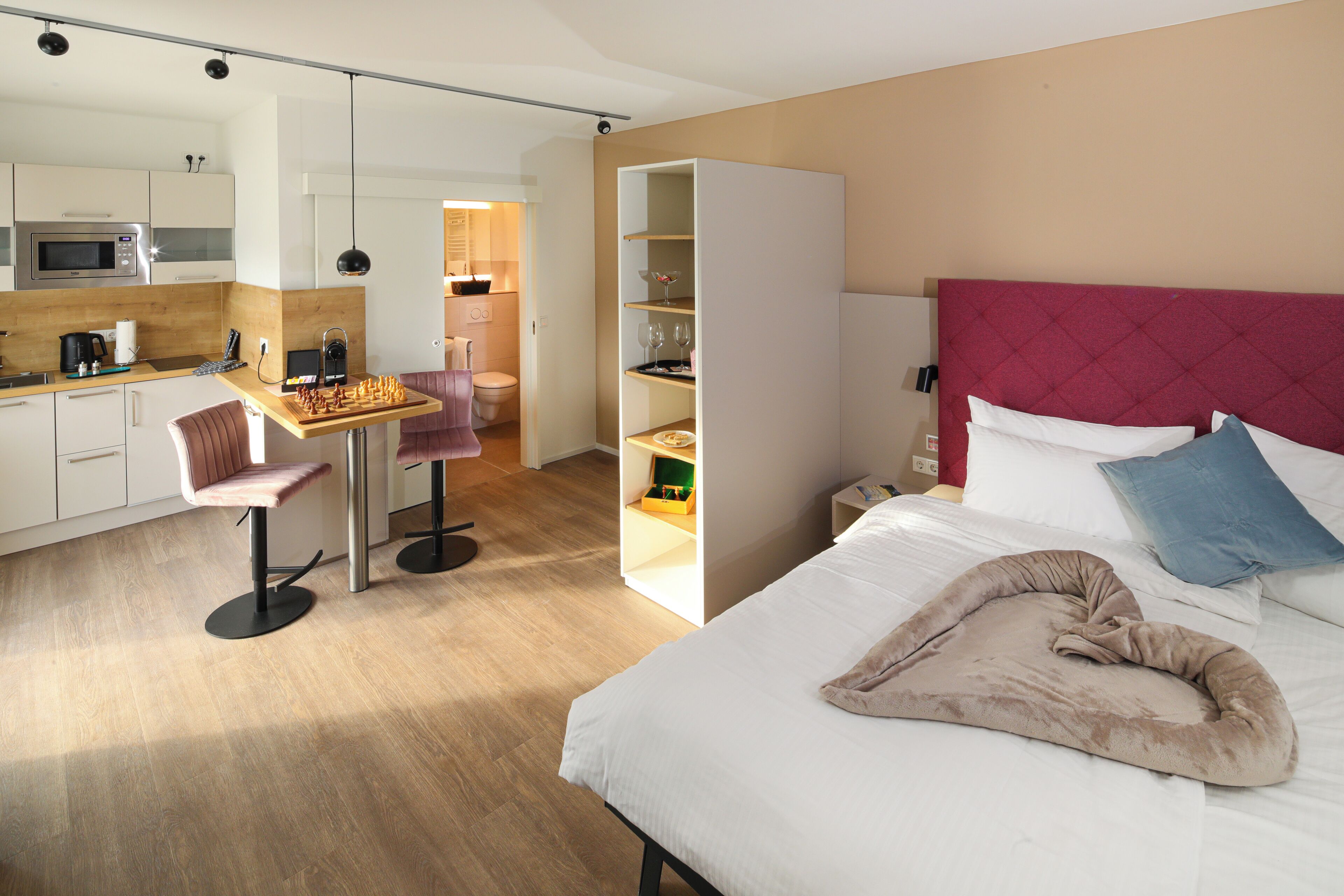 Una habitación de hotel compacta con kitchenette, zona de comedor y cama con cabecera roja.