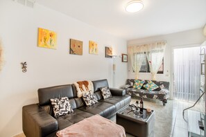 A living room with a sofa, a coffee table, and a TV. There are paintings on the wall and a window with curtains.