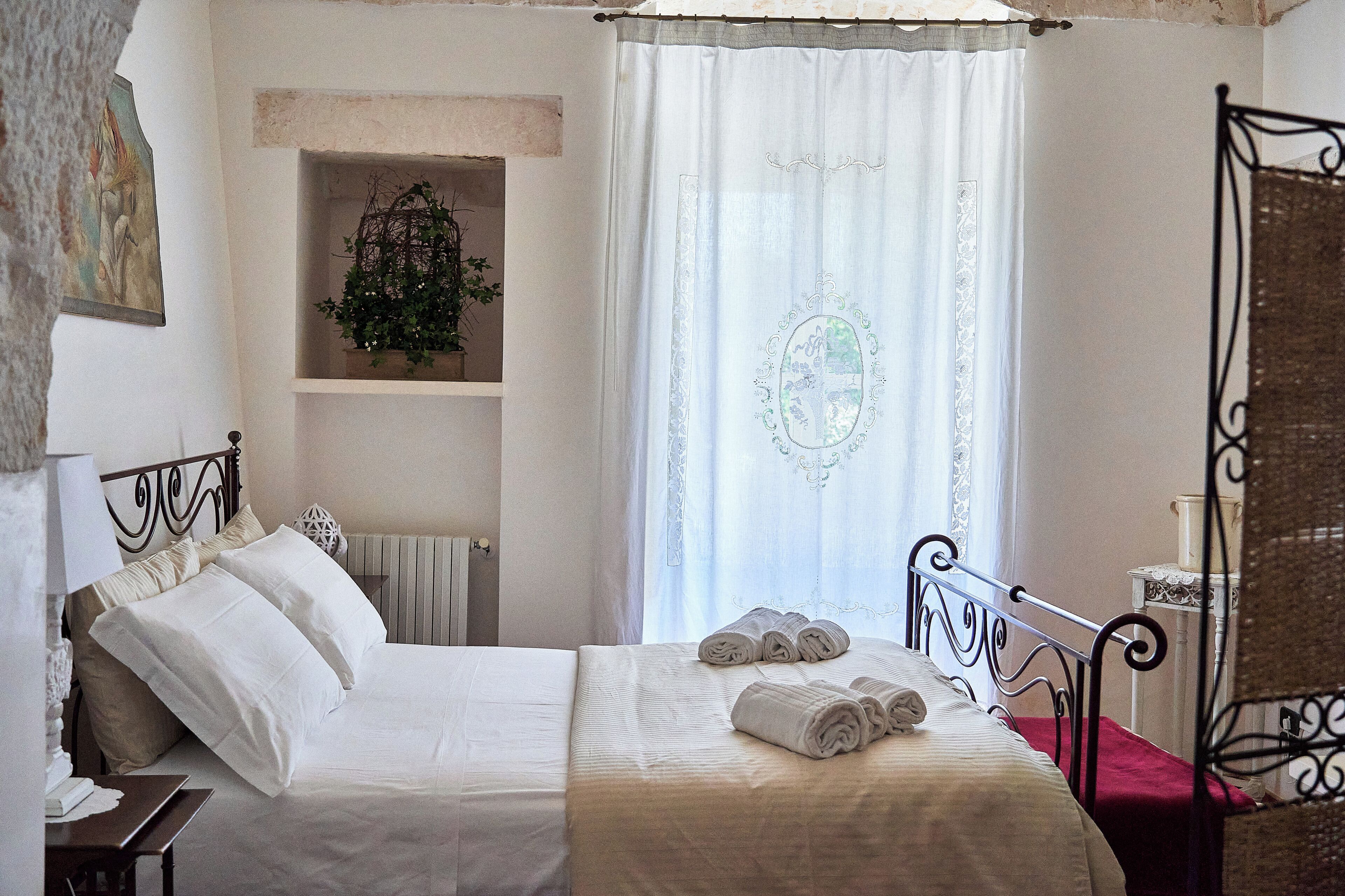 A bedroom with a bed, pillows, a bedside table, a window with curtains, a plant on a shelf, and a chair.
