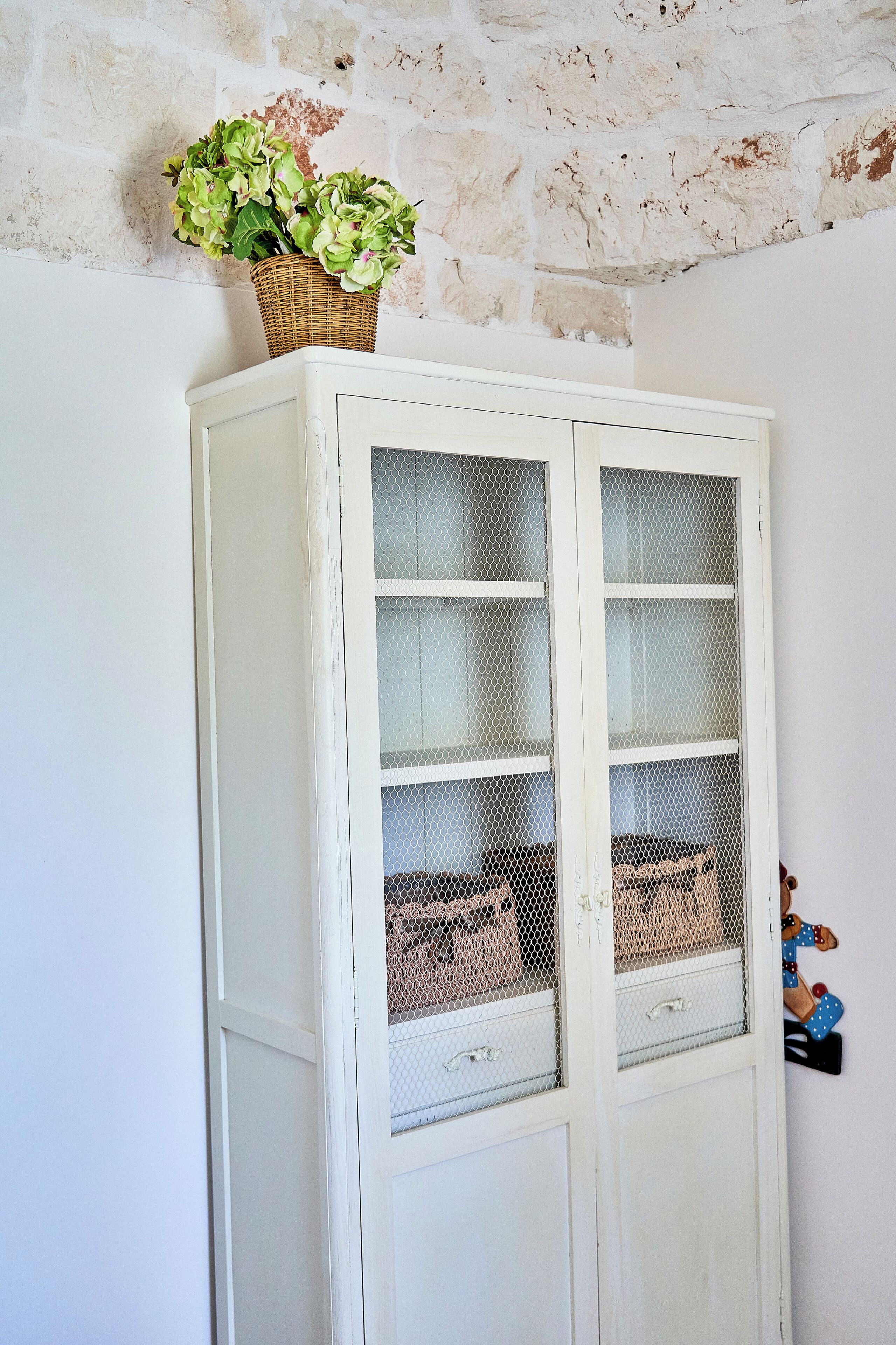 Un mueble blanco con puertas de vidrio, una cesta de mimbre encima y una pequeña figurina en la pared.