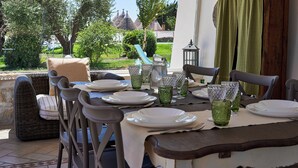 Uma área de refeições ao ar livre com móveis de vime, uma mesa posta para quatro pessoas e vista para um jardim e edifícios ao fundo.