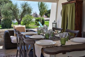 Ein Essbereich im Freien mit Korbmöbeln, einem Tisch für vier Personen und Blick auf einen Garten sowie Gebäude im Hintergrund.