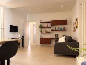 Un salon moderne comprenant un canapé, un téléviseur fixé au mur, une bibliothèque et un coin repas avec des chaises.