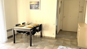 A dining area with a table set for four, chairs, a painting on the wall, and a door leading to another room. - Apartment "Bord de Mer" 5min beach, pool, AC (Fréjus)