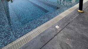 En swimmingpool med flisegange, omgivet af en betonflade, med høje bygninger i baggrunden.
