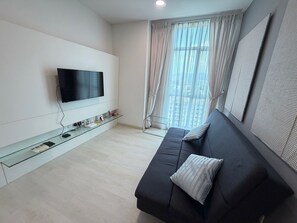 Un salon moderne doté d’un téléviseur à écran plat, d’une table basse en verre, d’un canapé foncé agrémenté d’un coussin et d’une grande fenêtre ornée de rideaux légers.